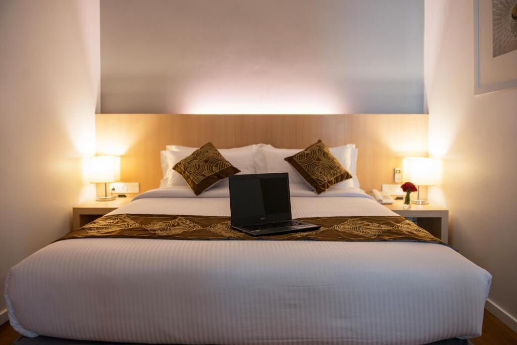 A hotel room with a large bed, two pillows, a laptop on the bed, bedside lamps, and a small table with a vase of flowers.