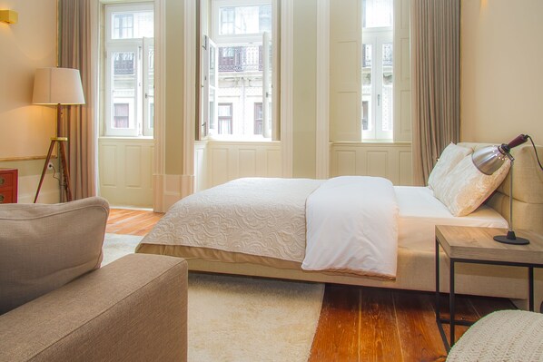 A hotel room with a large bed, a bedside table with a lamp, a chair, and a view of buildings through the windows. - Liiiving in Porto - Art & Heart Studio (Porto)