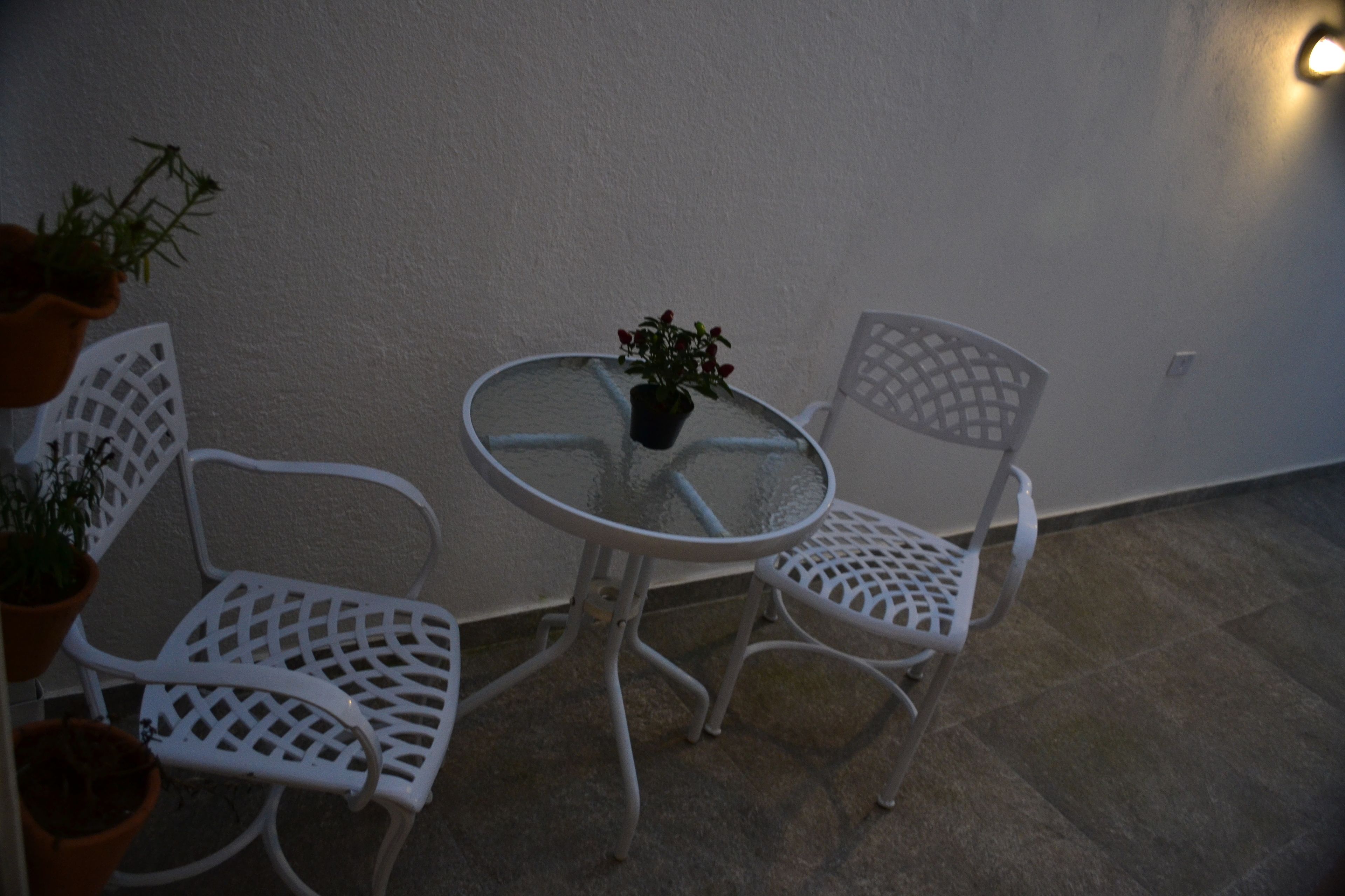 A small outdoor seating area with a round glass table and two white metal chairs. There are potted plants on the left side.