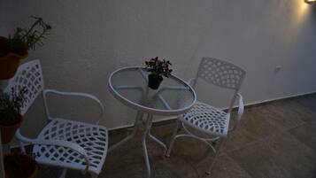 A small outdoor seating area with a round glass table and two white metal chairs. There are potted plants on the left side.