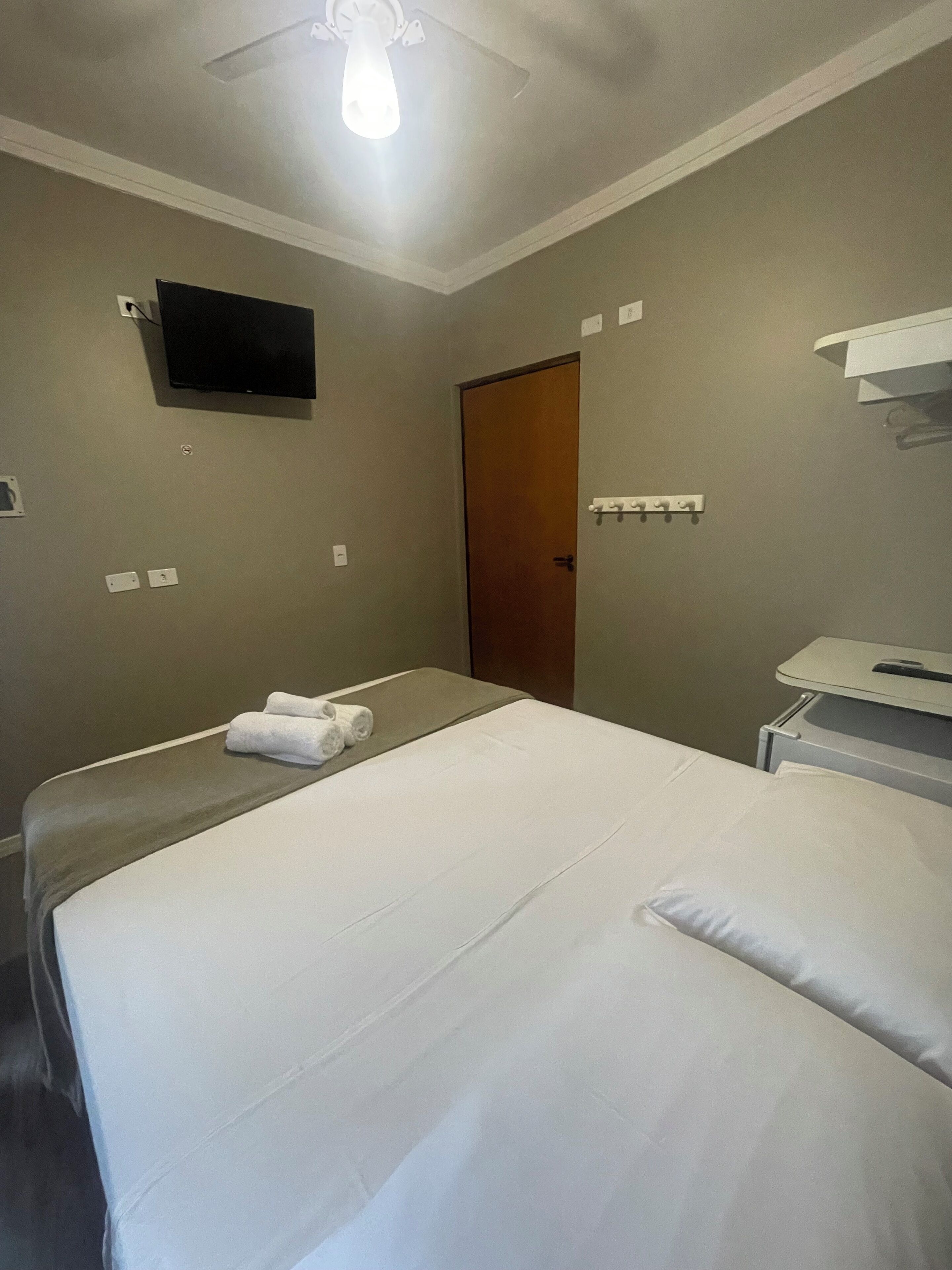 Quarto de hotel com duas camas, uma televisão de tela plana fixada na parede, uma porta de madeira e uma pequena mesa com um espelho.