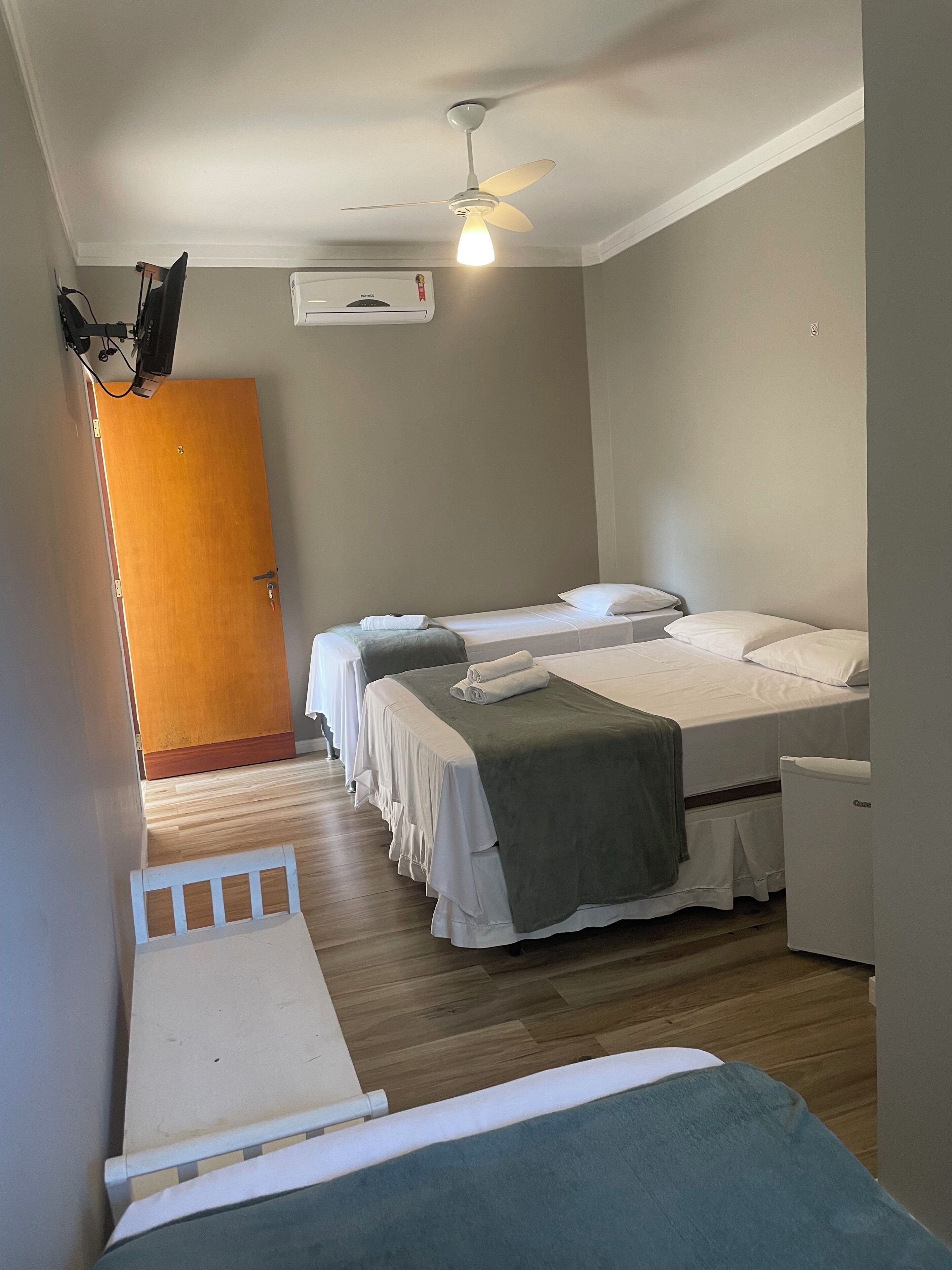 Quarto de hotel com duas camas, um ventilador de teto, um aparelho de ar condicionado e uma porta de madeira.