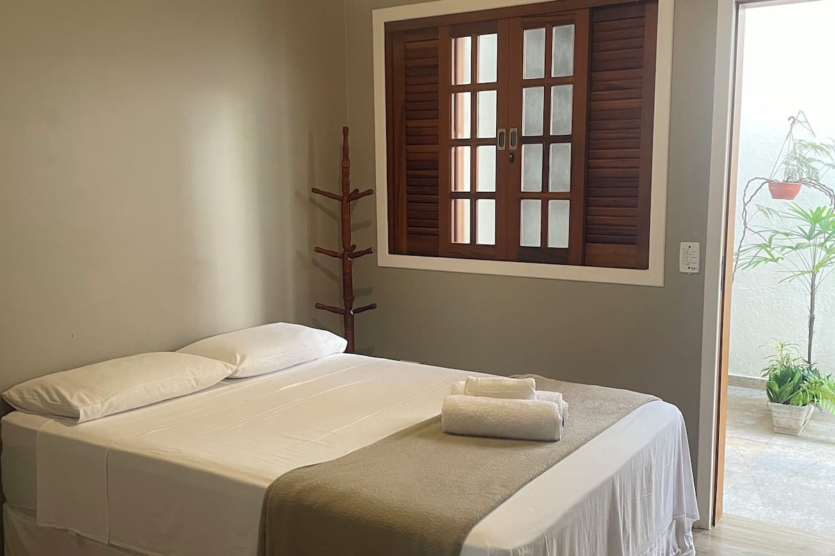 A neatly made bed with white linens and a beige throw blanket, a wooden coat rack, a ceiling fan, and an air conditioning unit.