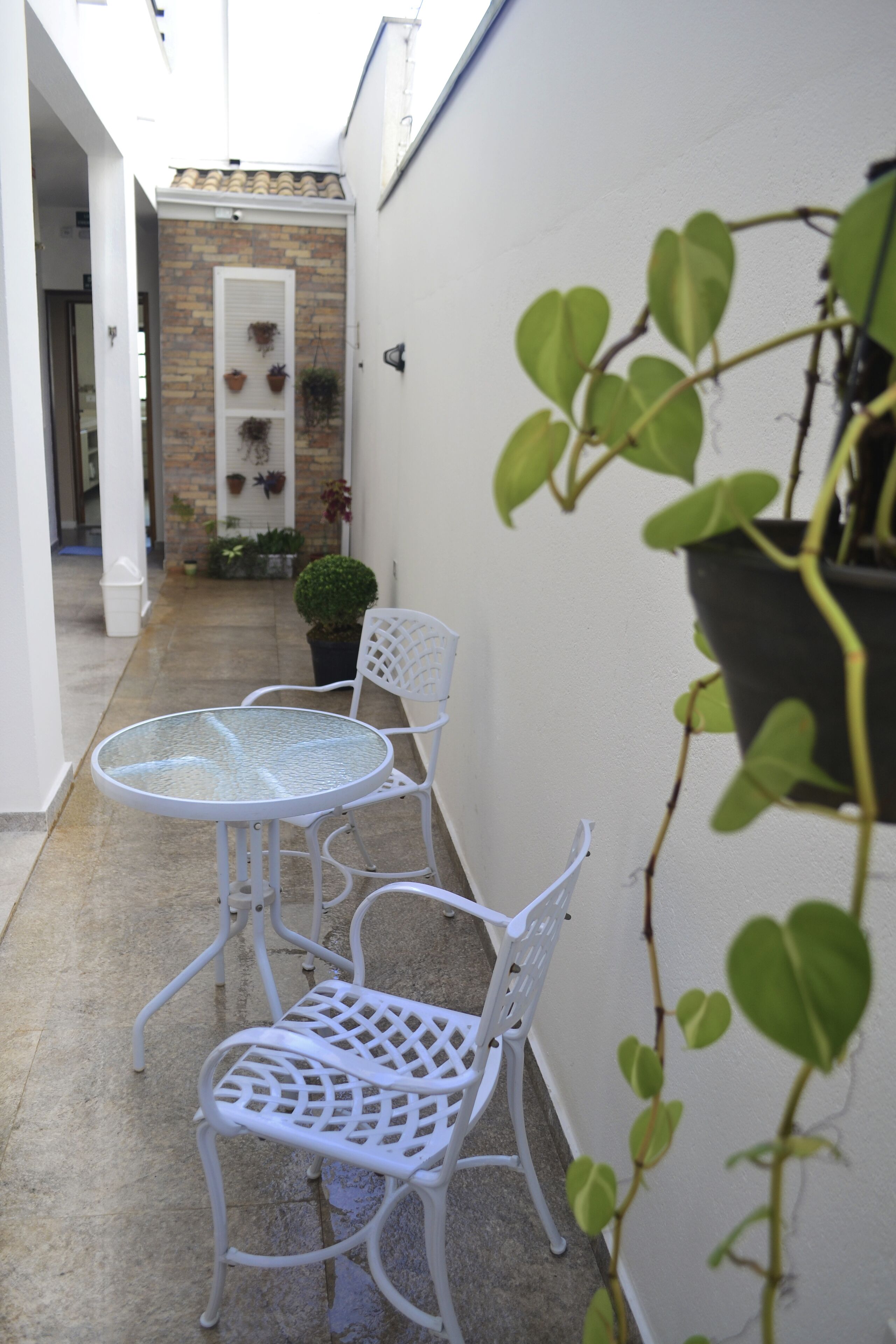 A narrow outdoor corridor with a glass table and white metal chairs, a potted plant, and a small tree.