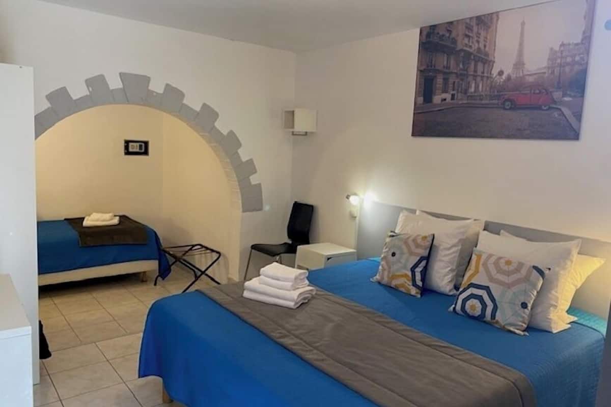 Ein Hotelzimmer mit einem großen Bett, einem kleineren Bett, einem Schreibtisch und einem Sessel. An der Wand hängt ein Gemälde und über der Tür befindet sich ein Schild.