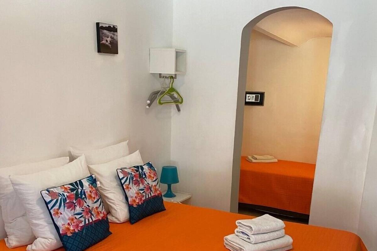 Ein Hotelzimmer mit einem großen Bett, weißen Kissen, einer orangefarbenen Tagesdecke, einer blauen Lampe, einem weißen Nachttisch, einem gerahmten Bild an der Wand und einem Spiegel, der das Bett reflektiert.