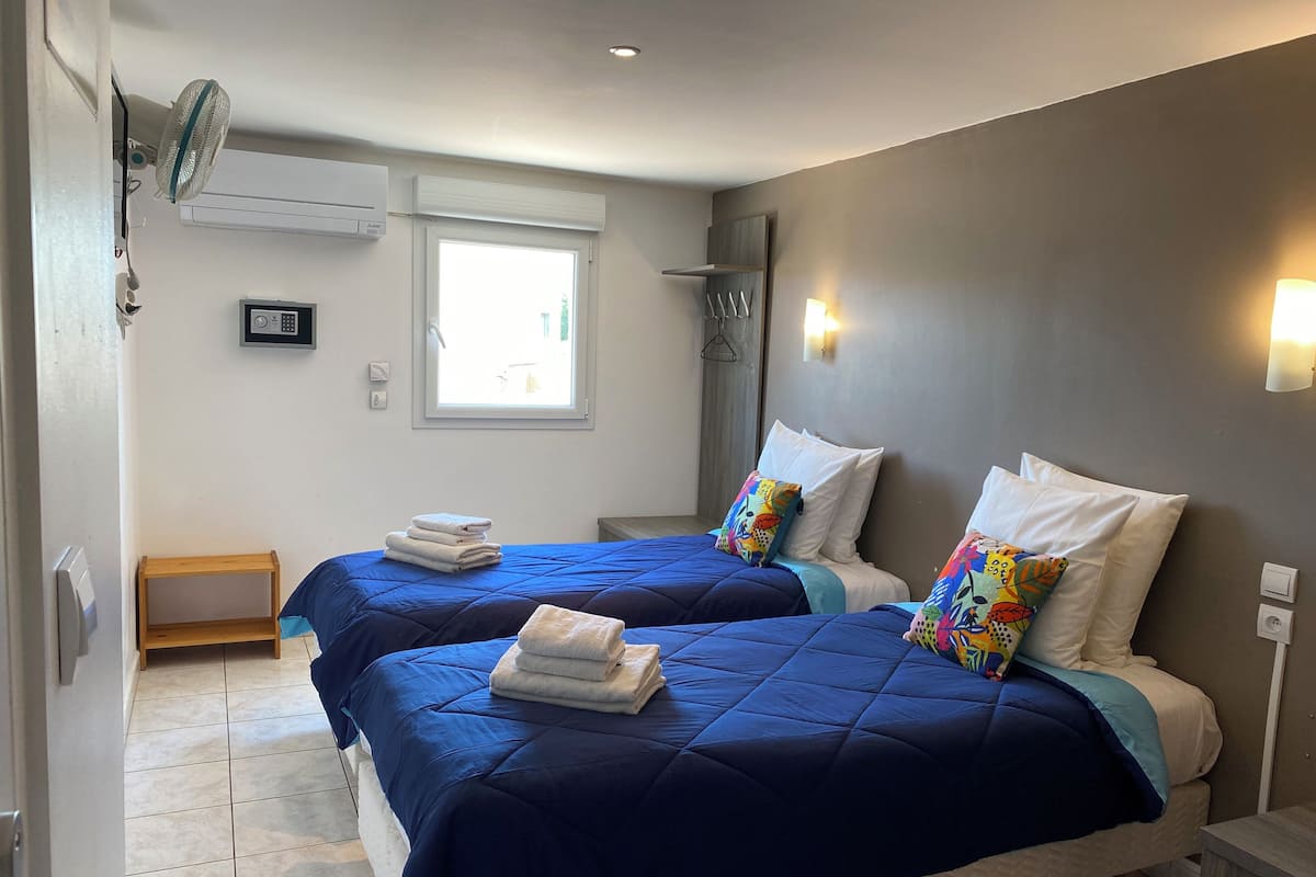 Ein Hotelzimmer mit zwei Betten, jeweils mit blauer Bettwäsche und weißen Kissen, einem kleinen Holfnachtkästchen, einem Standventilator und einem Fenster mit Blick ins Freie.
