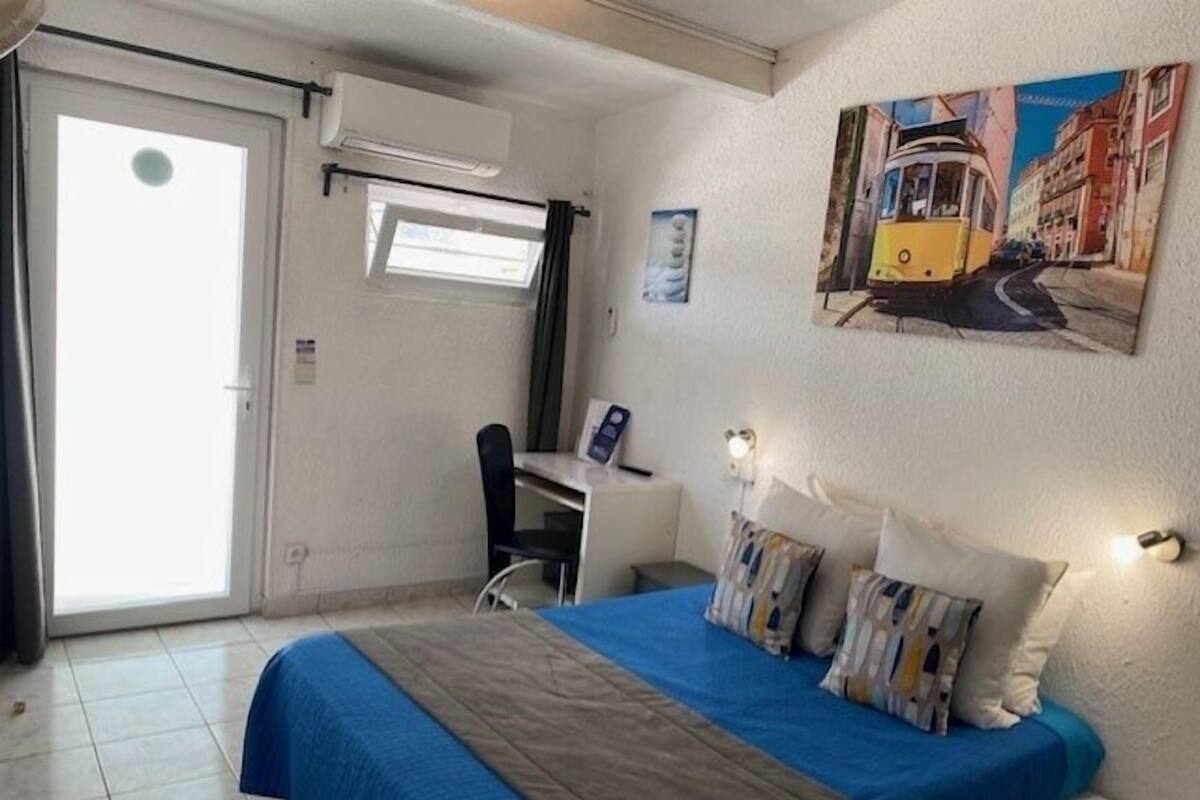Ein Hotelzimmer mit Bett, Schreibtisch und Stuhl. Es gibt ein Fenster mit Vorhängen, eine Wand-Klimaanlage und ein Gemälde eines Straßenbahns.
