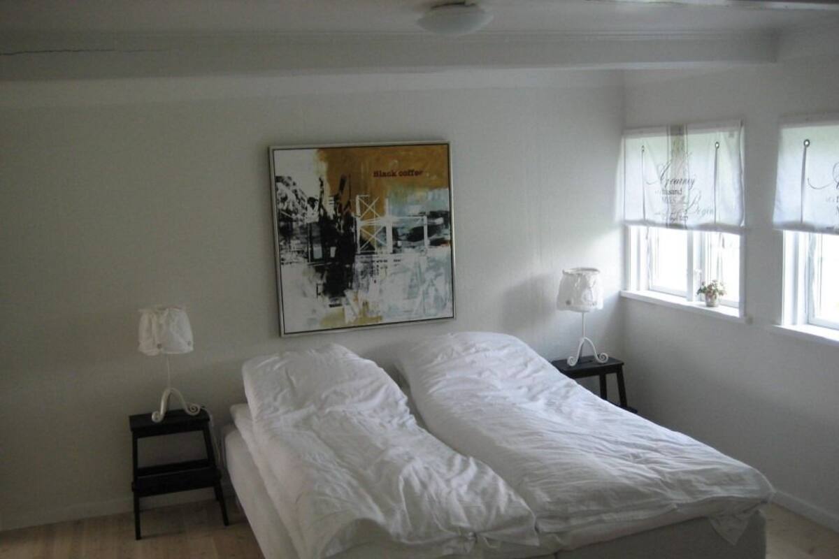 A bedroom with a bed, a nightstand, a lamp, a window with curtains, and a wall-mounted picture.