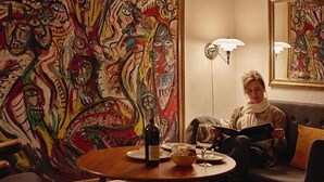 Iemand zit op een bank in een kamer met een kleurrijk abstract schilderij aan de muur, een klein rond tafeltje met een fles en glazen, en twee wandlampen.