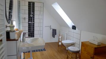 Un baño moderno con una bañera independiente, un lavabo e inodoro. Hay un tragaluz sobre el inodoro, y un taburete de madera con cojín colocado a su lado.