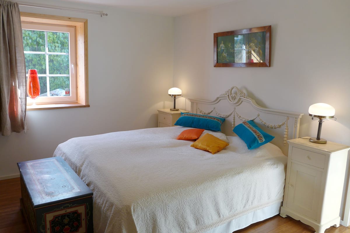 Ein ordentlich eingerichtetes Schlafzimmer mit Bett, Nachttischen, einem Fenster mit Vorhängen und einem gerahmten Bild an der Wand.