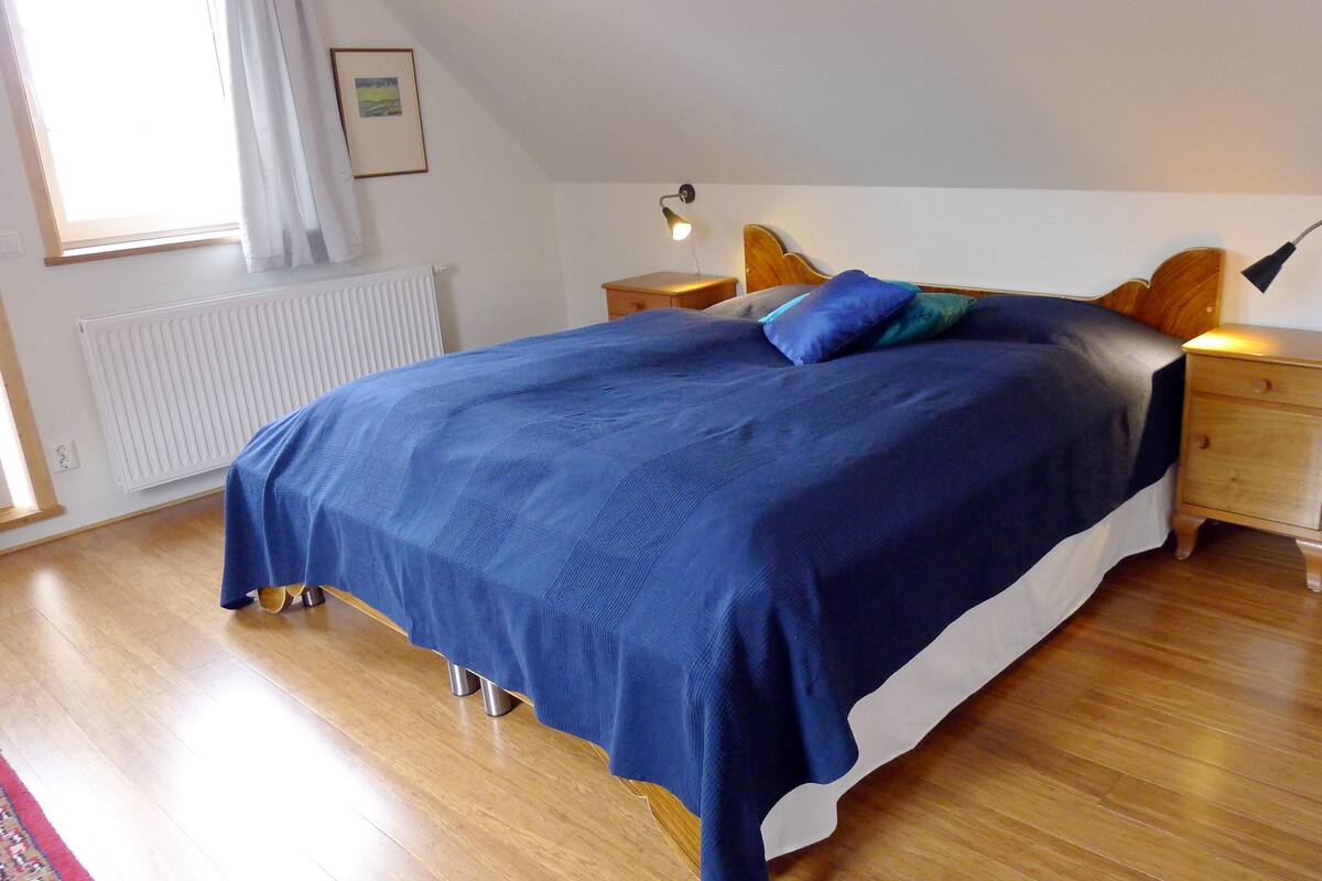 Ein Schlafzimmer mit einem großen Bett, Holz-Nachttischen, einem Beistelltisch, einer Heizung und einem Fenster mit Vorhängen.