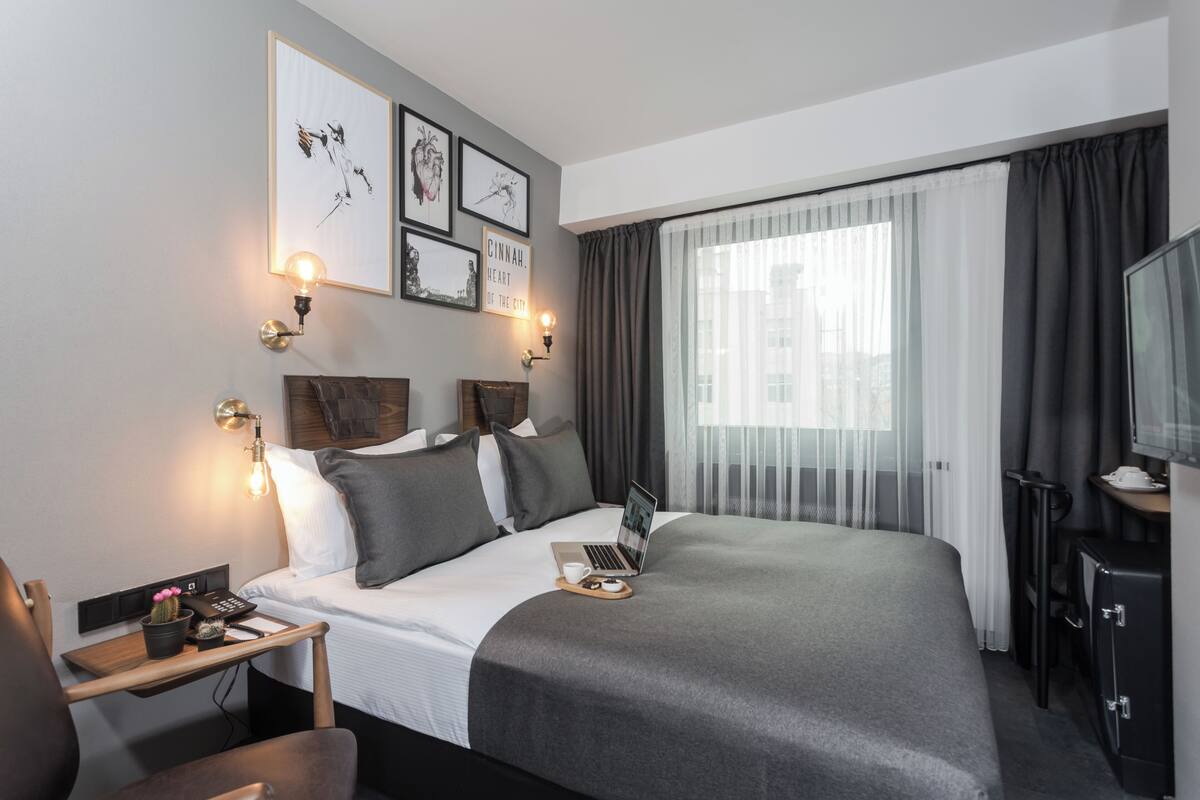 A modern hotel room with a large bed, a desk with a laptop, a TV, and artwork on the wall.