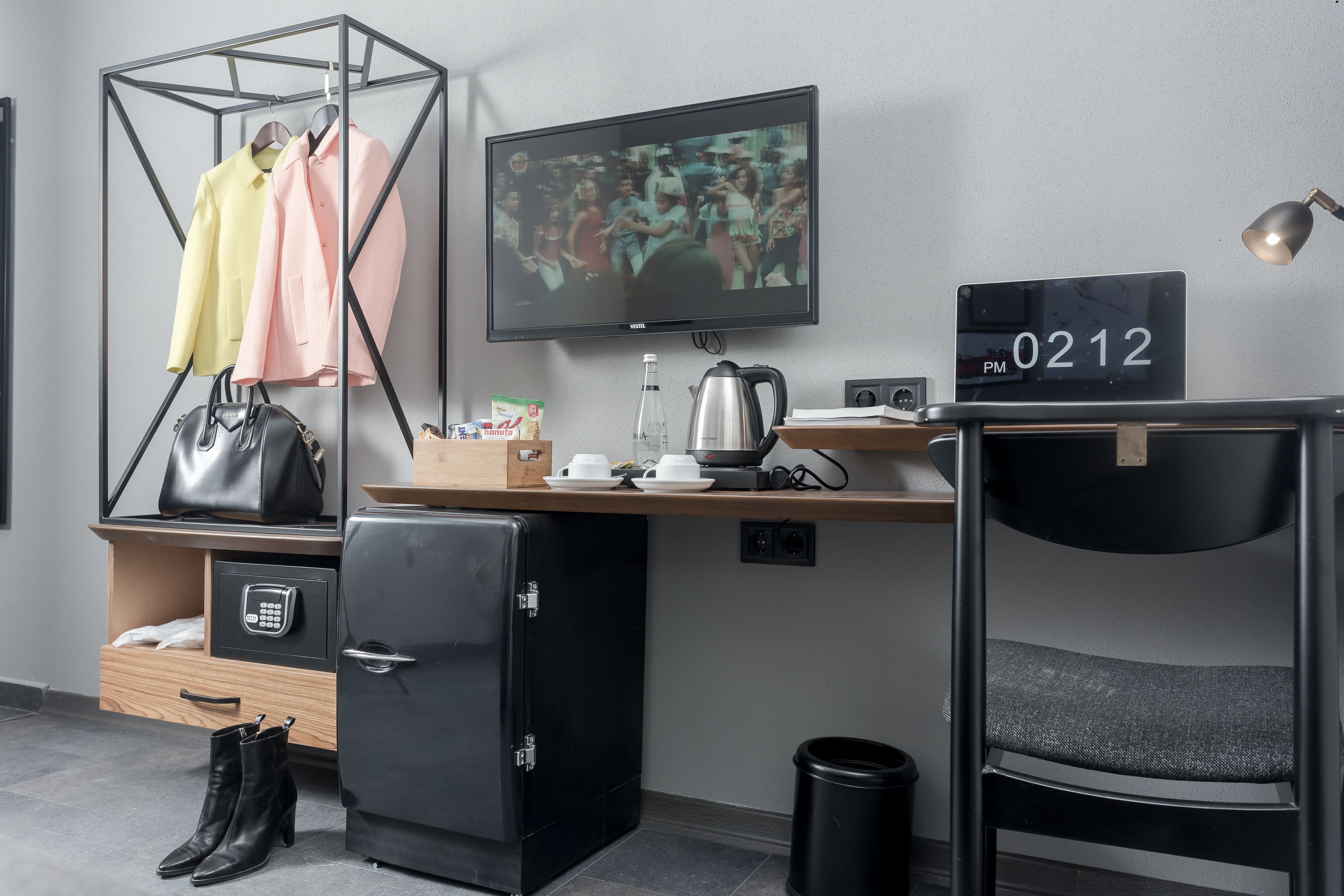Une chambre d’hôtel compacte comprenant un coin cuisine, une télévision, un petit réfrigérateur, un bureau avec un ordinateur, une chaise et un porte-manteau mural.