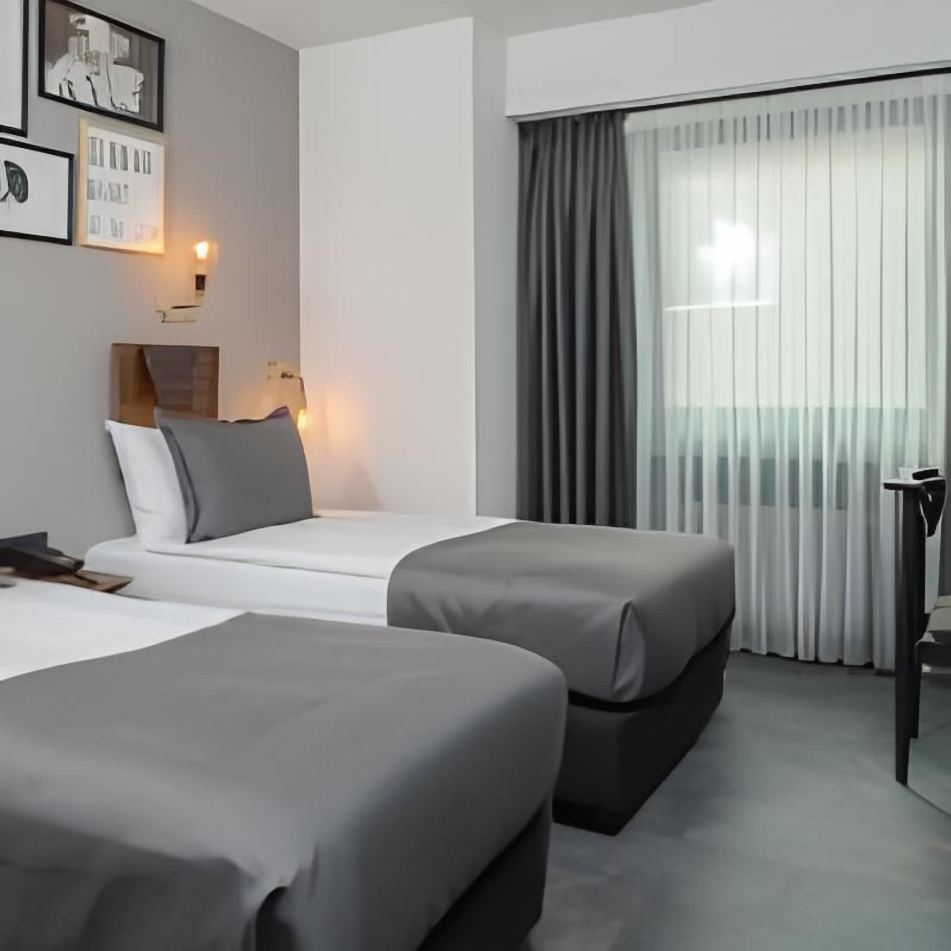Una habitación de hotel con dos camas, una mesita de noche, una lámpara y un ventanal con cortinas.