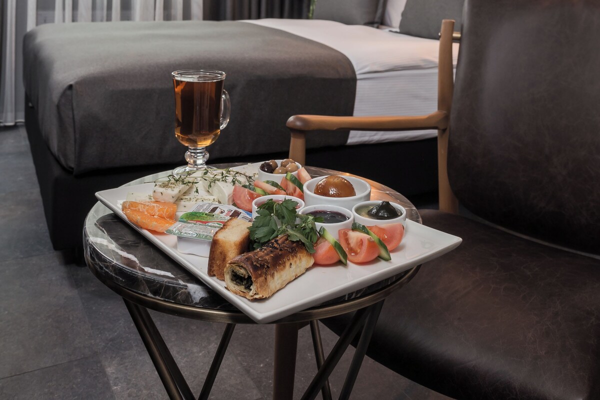 A hotel room with a bed, a dining table with food, a chair, and artwork on the wall.