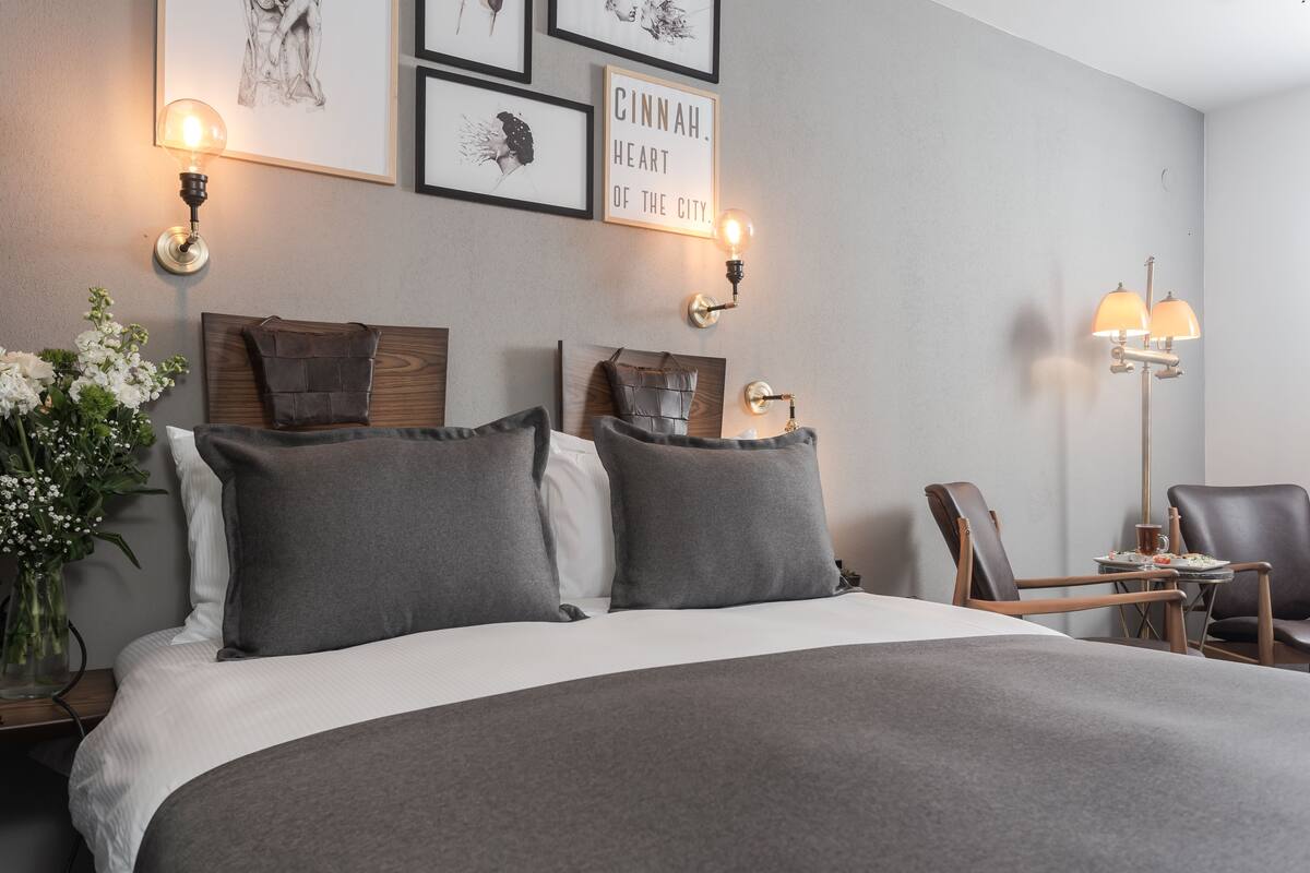 A hotel room with a large bed, two wall-mounted lamps, a framed quote, a vase with flowers, and a small table with a chair.