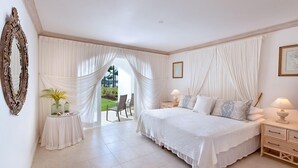 A bedroom with a large bed, two bedside tables with lamps, a mirror, and a view of a garden through sheer curtains.