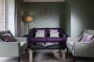 Una sala moderna con un sofá morado, dos sillones, una mesa de centro y un adorno geométrico en la pared.