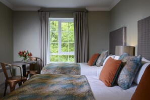 A hotel room with two beds, a window with curtains, a table with flowers, and a lamp. - The Reserve at Muckross (Killarney)