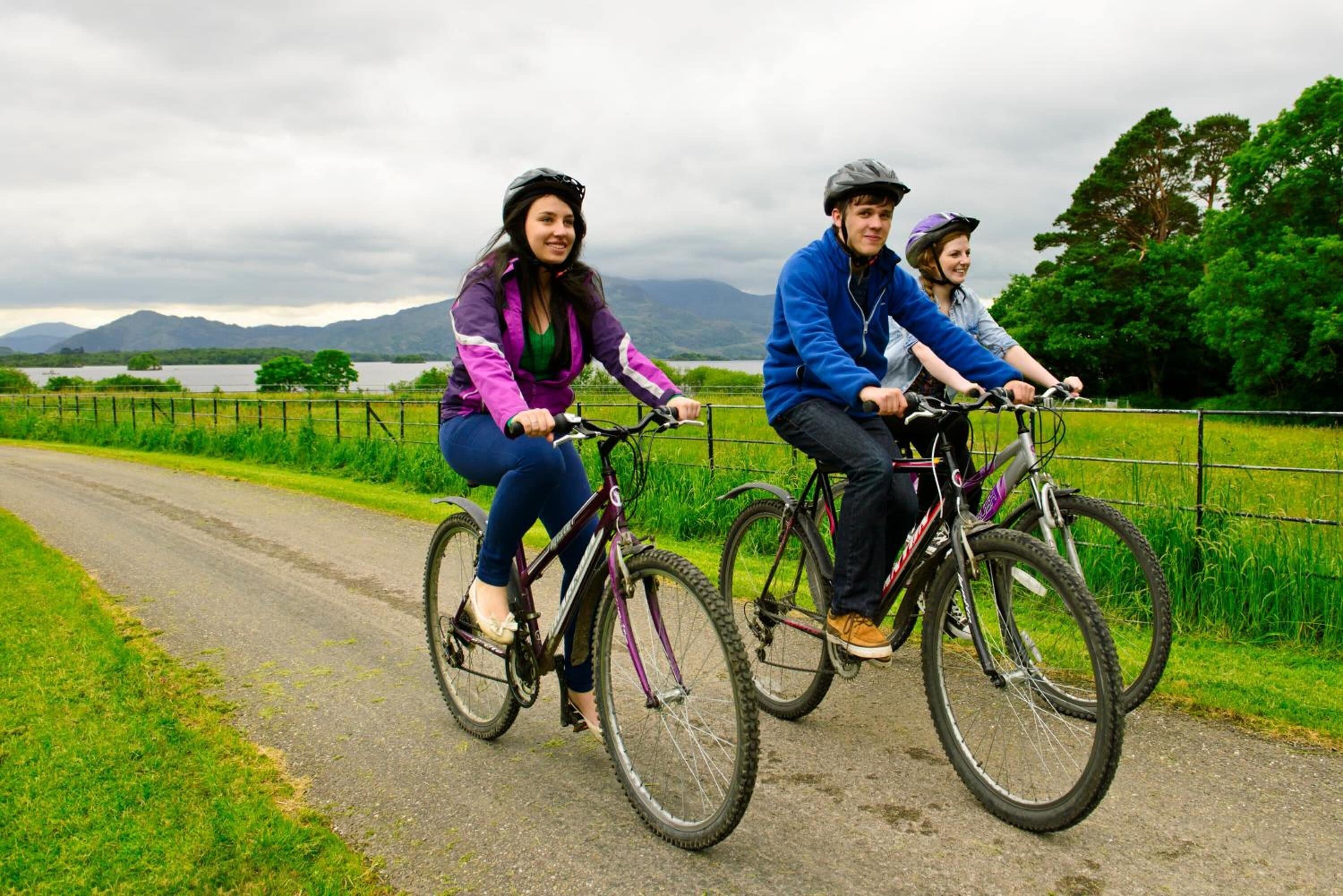 Three people riding bicycles on a rural road with a lake and mountains in the background.