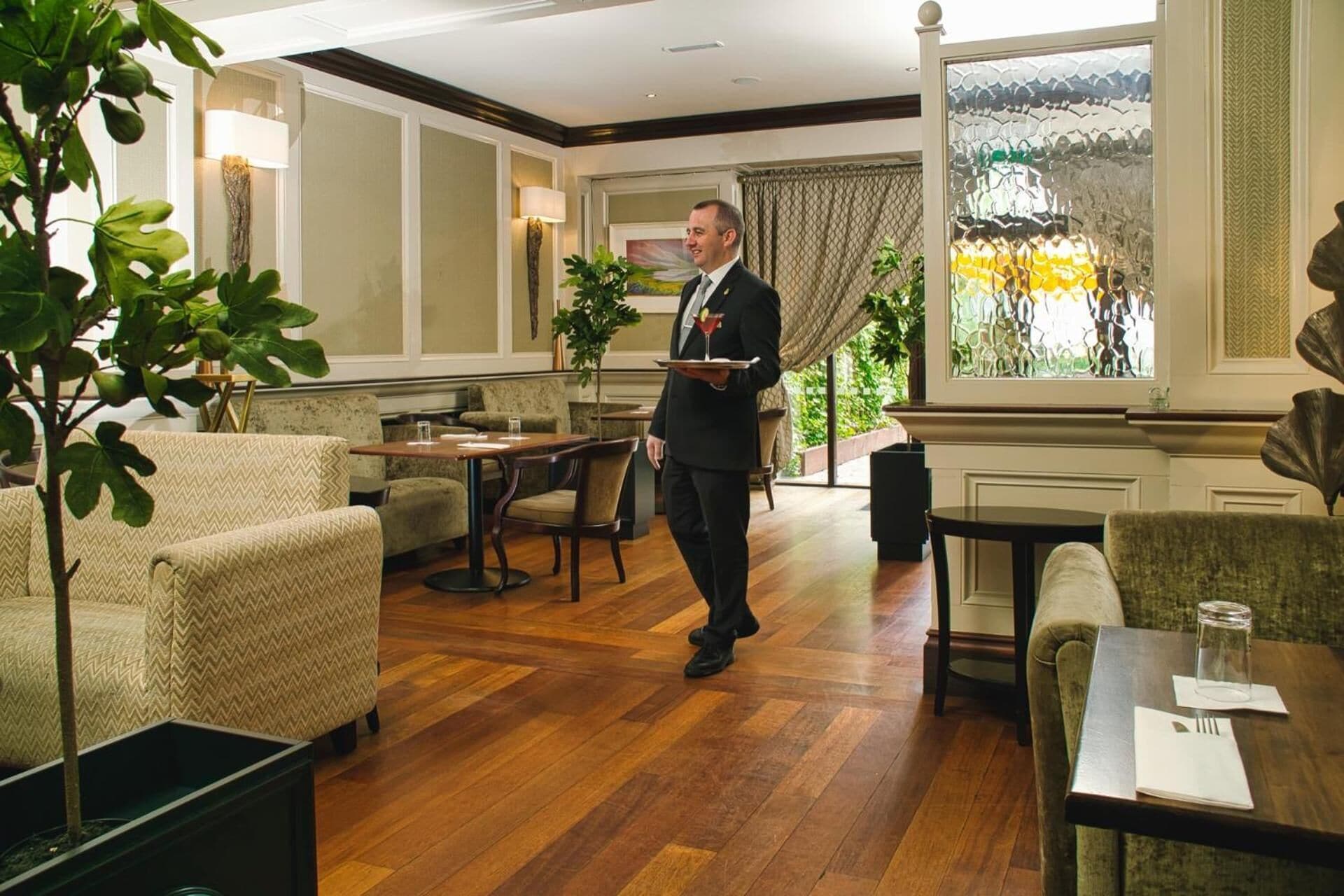 A hotel lobby with wooden flooring, upholstered seating, a waiter carrying a tray, and a large window with a view.