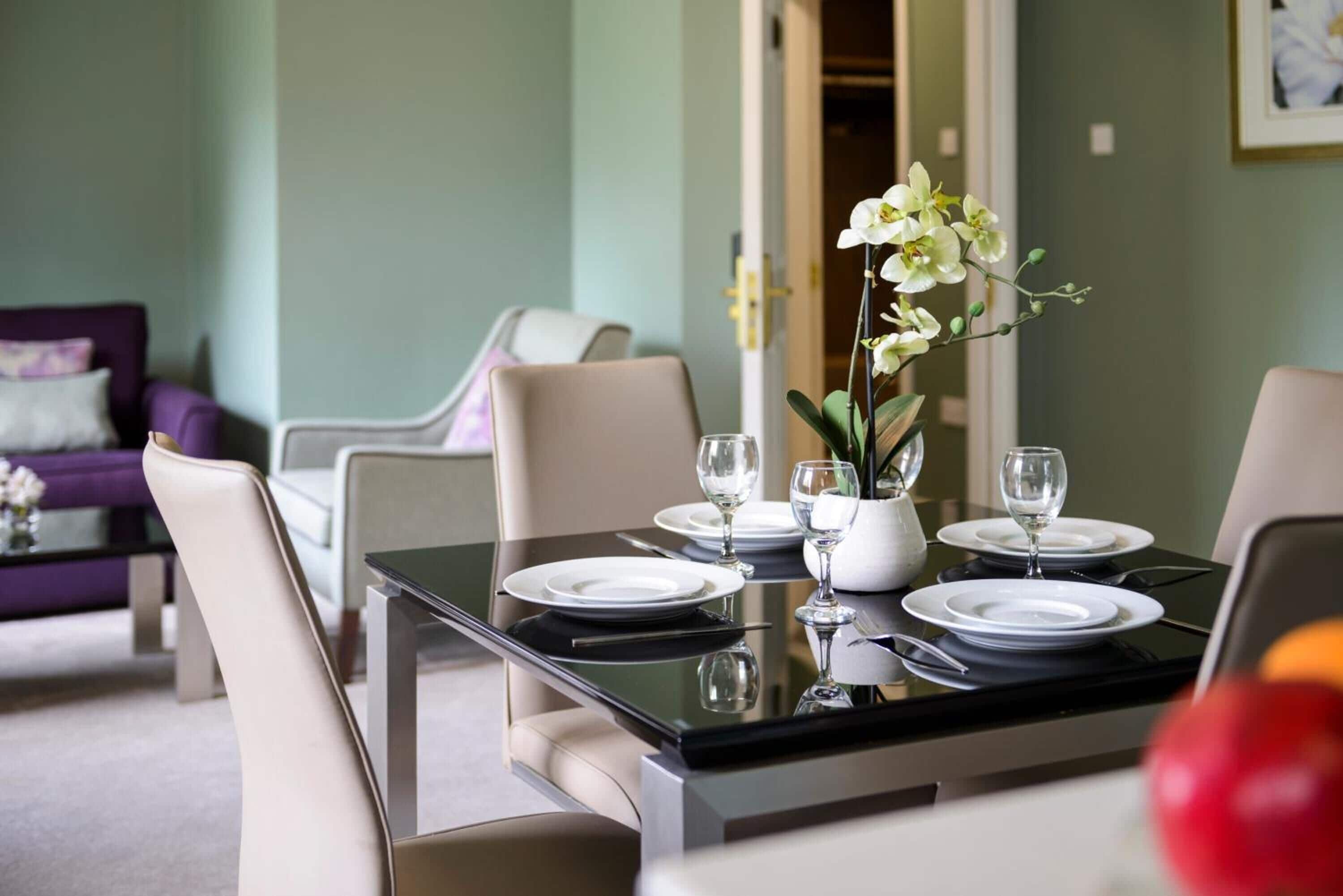 A dining table set with white plates, glasses, and a vase with white flowers.