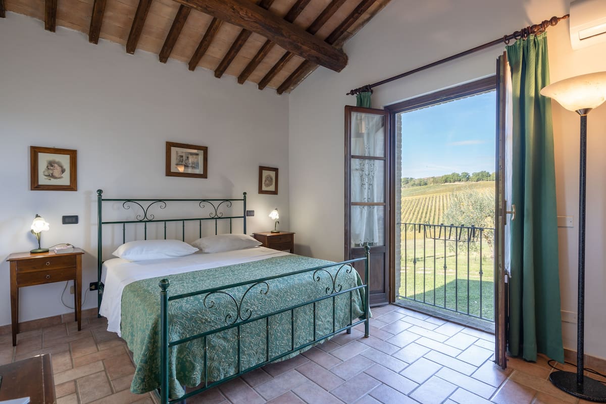 A bedroom with a metal bed frame, a green bedspread, a nightstand with a lamp, and a view of a green field through a glass door.