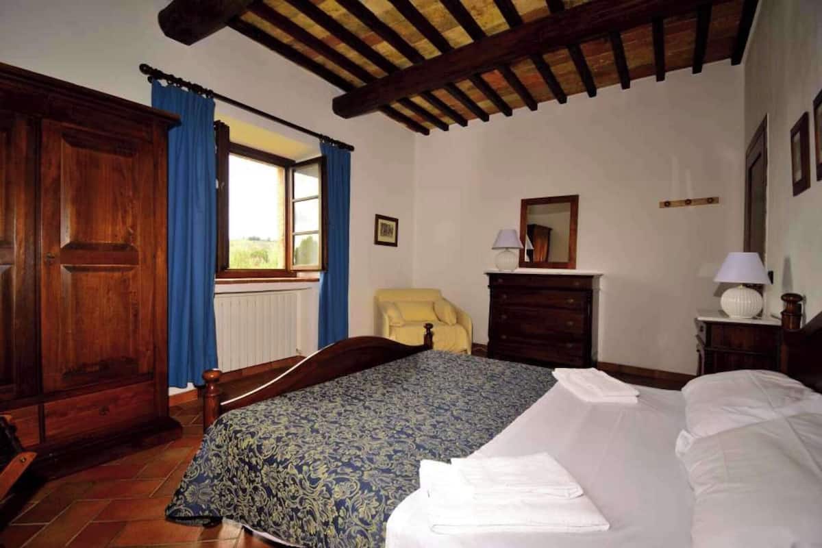 A bedroom with a bed, a wooden wardrobe, a chair, a dresser, a window with curtains, and a ceiling with exposed beams.