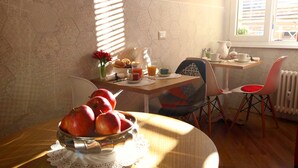 A dining area with a wooden table, a bowl of apples, and a pitcher of orange juice. There are chairs and a window with blinds. - Campanella3 (Rome)