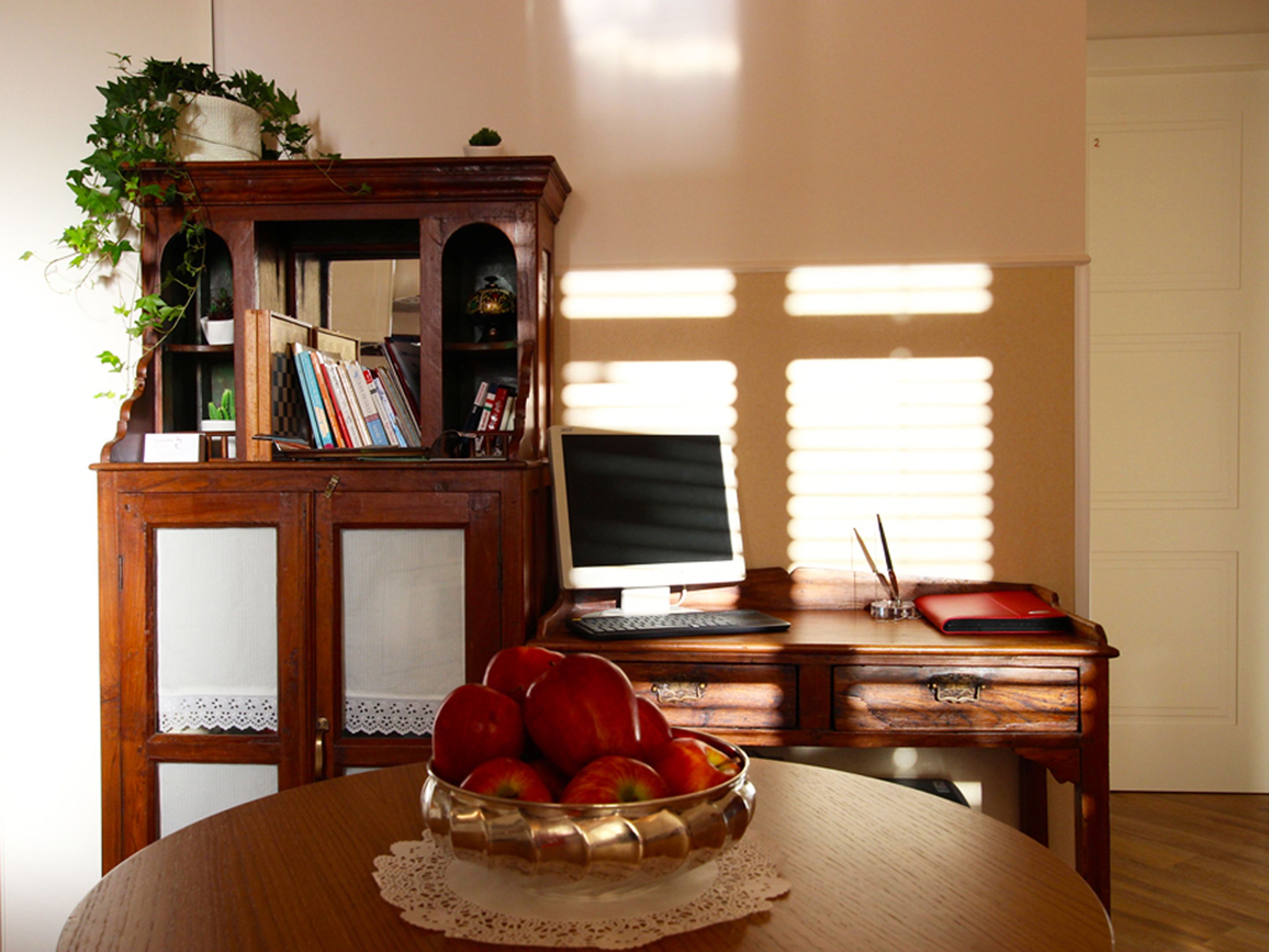 A wooden desk with a computer, a bowl of apples, and a bookshelf with books and plants.