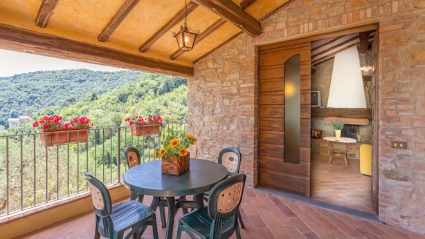 Ein Balkon mit Steinmauer, Holzboden und einem Tisch mit Stühlen. Auf der Brüstung stehen Blumentöpfe und es bietet sich ein Blick auf eine grüne Landschaft.