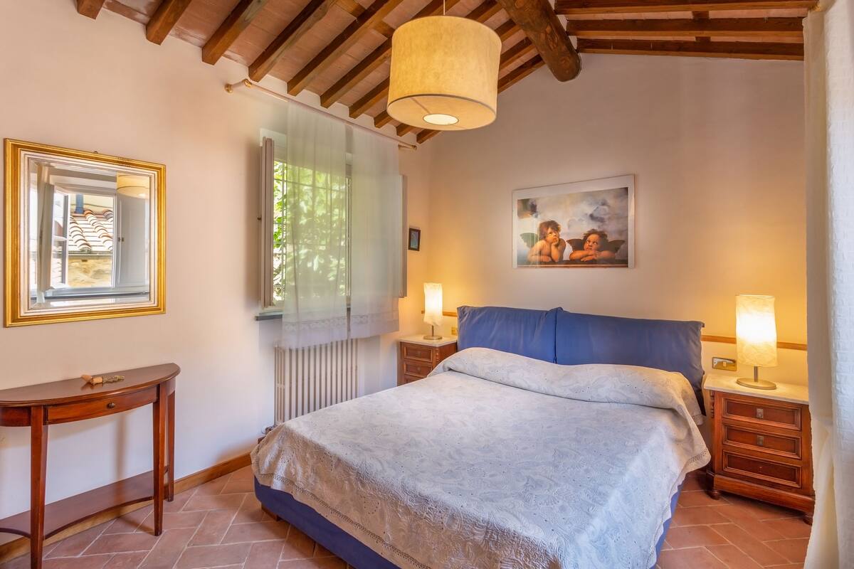A bedroom with a bed, bedside lamps, a wooden nightstand, a small table, a framed picture, and a window with a view of greenery.