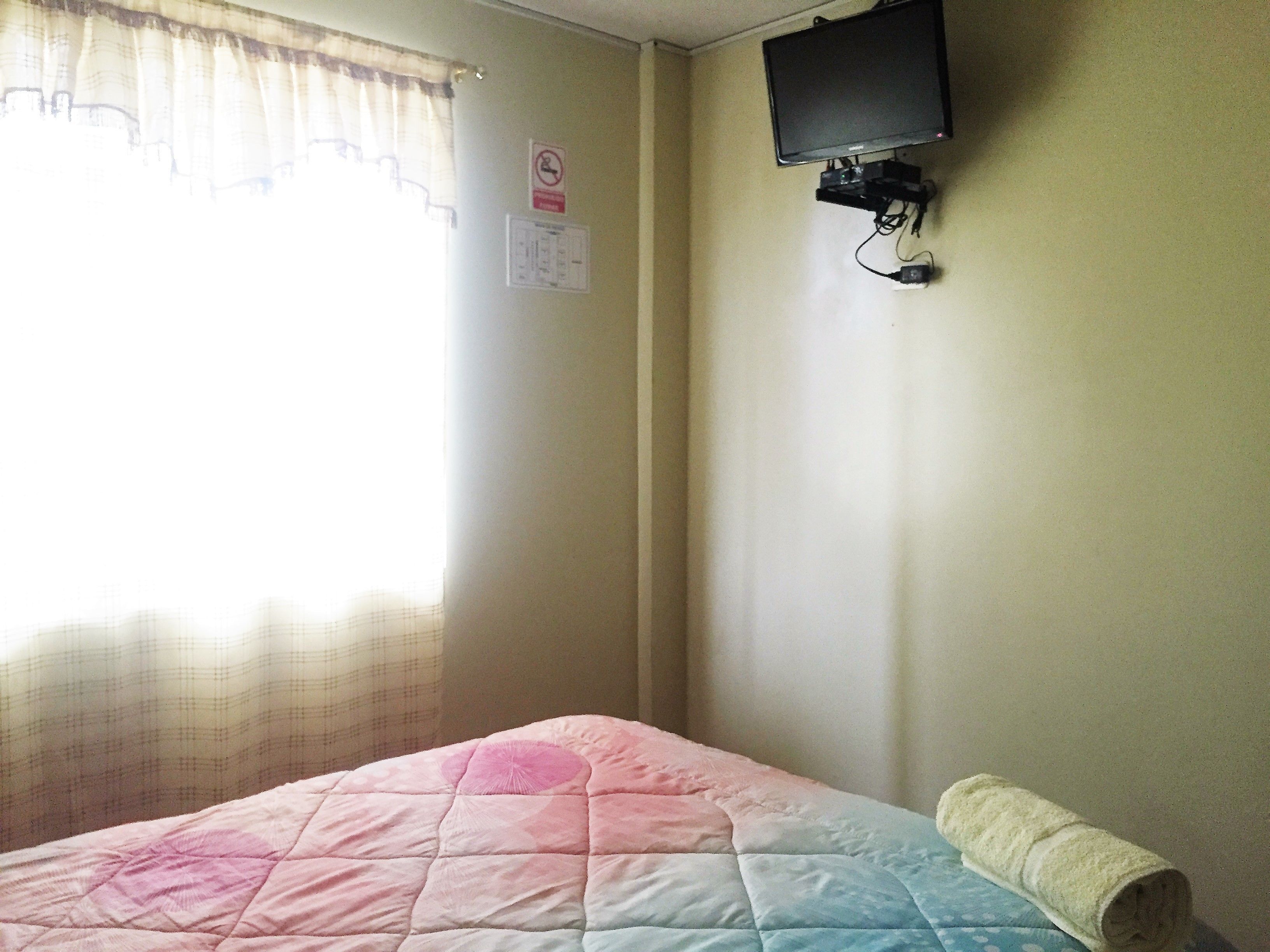 Une chambre avec un lit, un téléviseur fixé au mur et une fenêtre avec des rideaux.