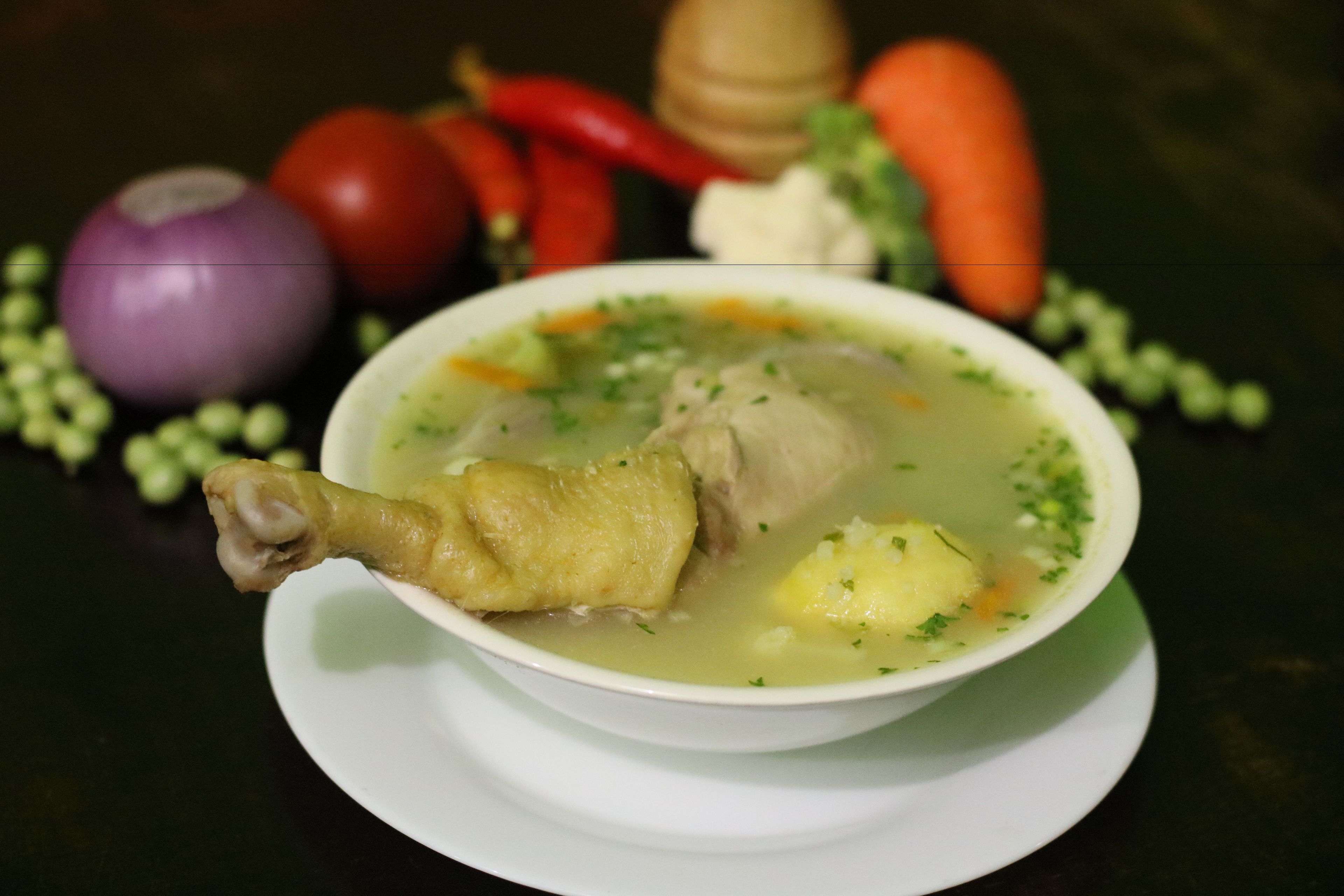 A bowl of soup with a chicken leg, carrots, and potatoes, surrounded by vegetables like tomatoes, onions, and peppers.