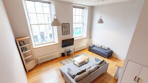 A modern living room with a grey sofa, a coffee table, and a bookshelf. There are large windows providing natural light and a view of the city. - Destiny Scotland - Broughton St Lofts (Edinburgh)