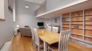 A modern dining area with a wooden table and white chairs, a sofa, and a built-in bookshelf. - Destiny Scotland - Broughton St Lofts (Edinburgh)
