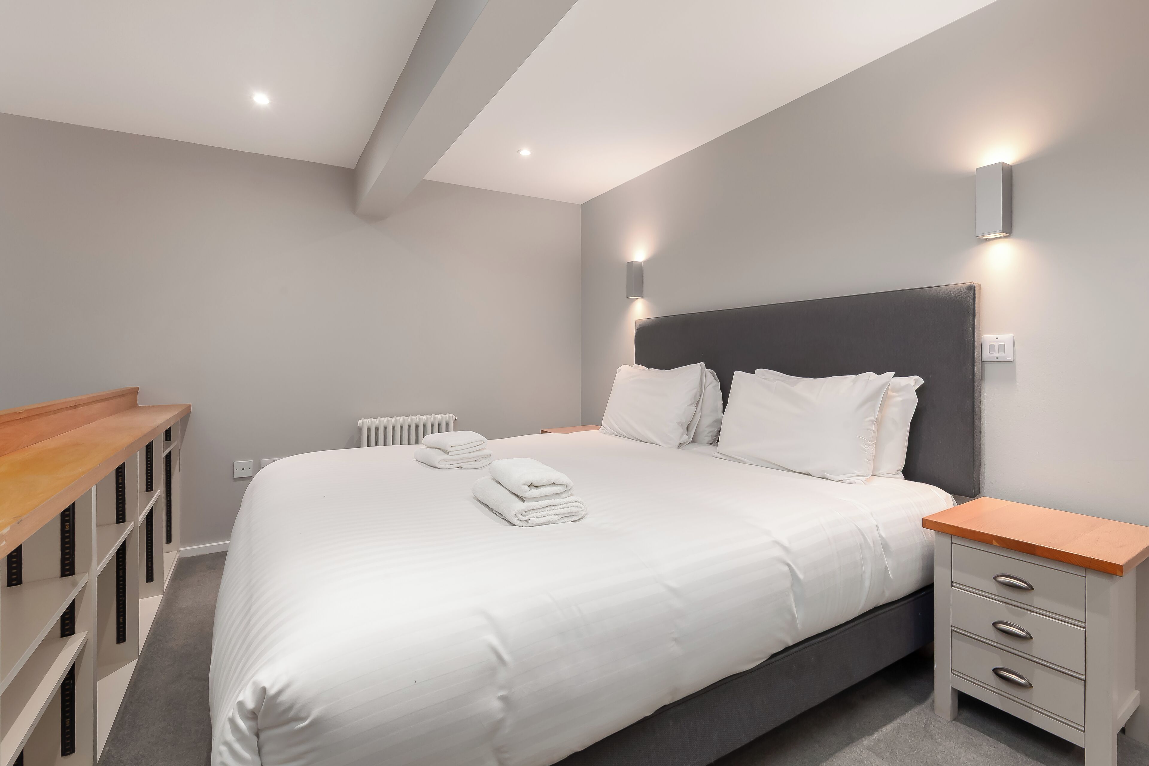 A neatly made bed with white linens and pillows, a bedside table with drawers, and a built-in shelving unit along the wall.
