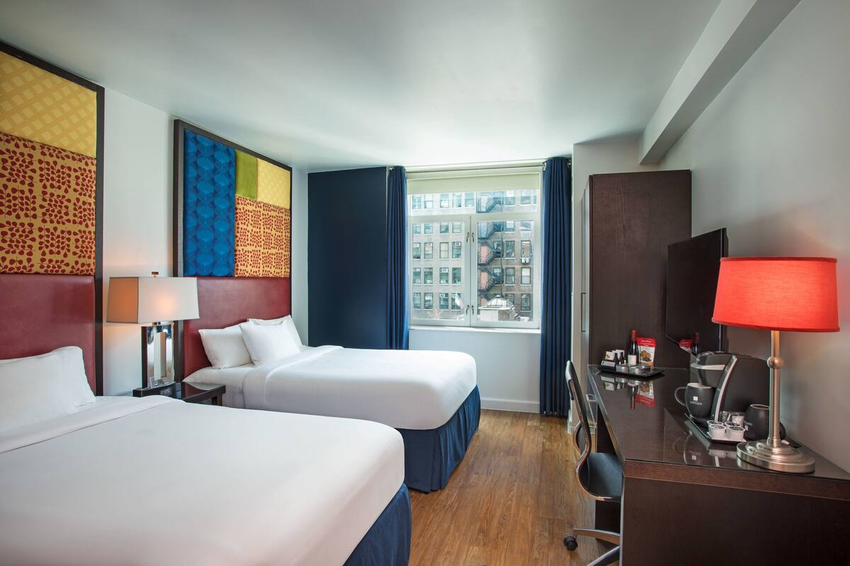 A hotel room with two beds, a desk with a lamp, a window with curtains, and a city view.