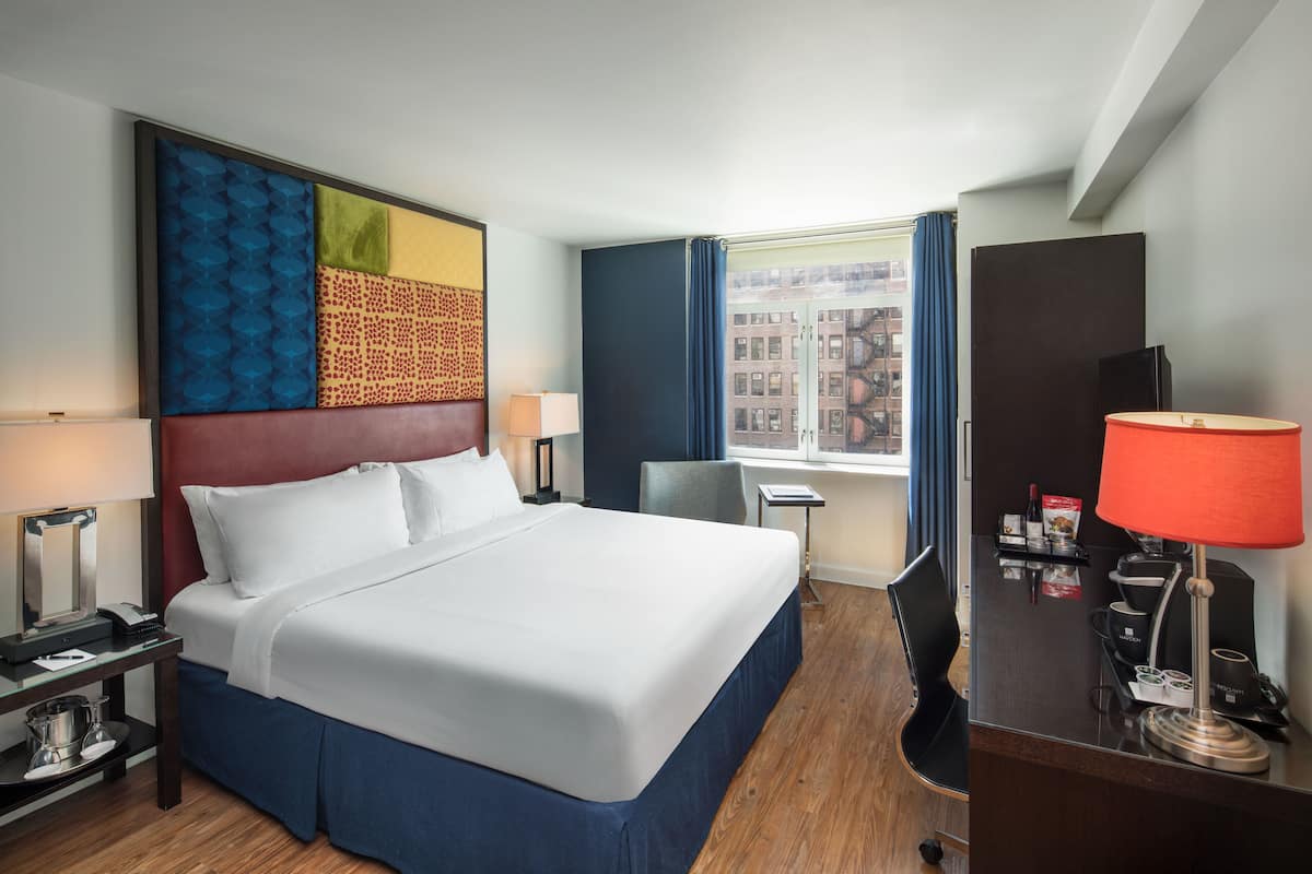 A hotel room with a large bed, a desk with a lamp, a coffee machine, and a view of buildings through the window.