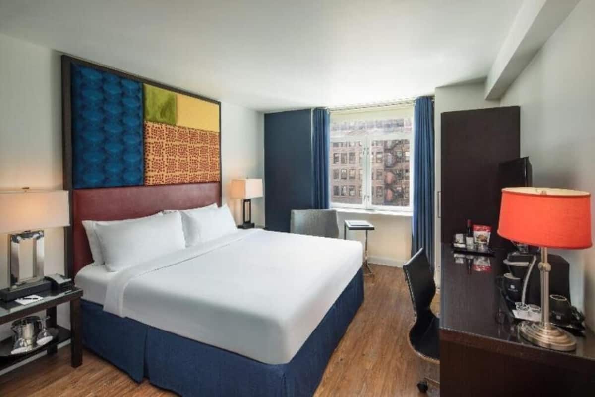 A hotel room with a bed, a desk with a lamp, a chair, a window with curtains, and a view of buildings.