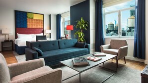 Moderni hotellihuone, jossa on suuri sänky, sohva, sohvapöytä ja suuret ikkunat, joista avautuu näkymä kaupungin ylle.