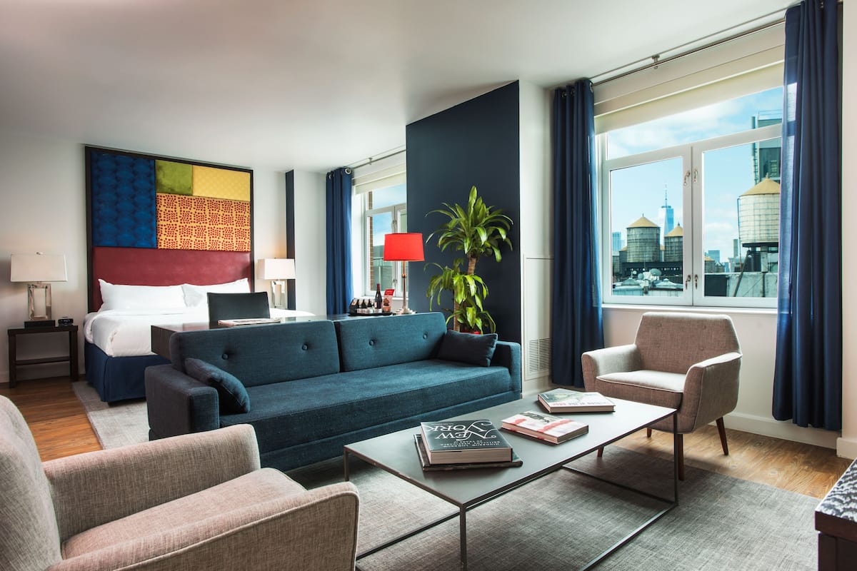 A modern hotel room with a large bed, a sofa, a coffee table, and a view of the cityscape through large windows.
