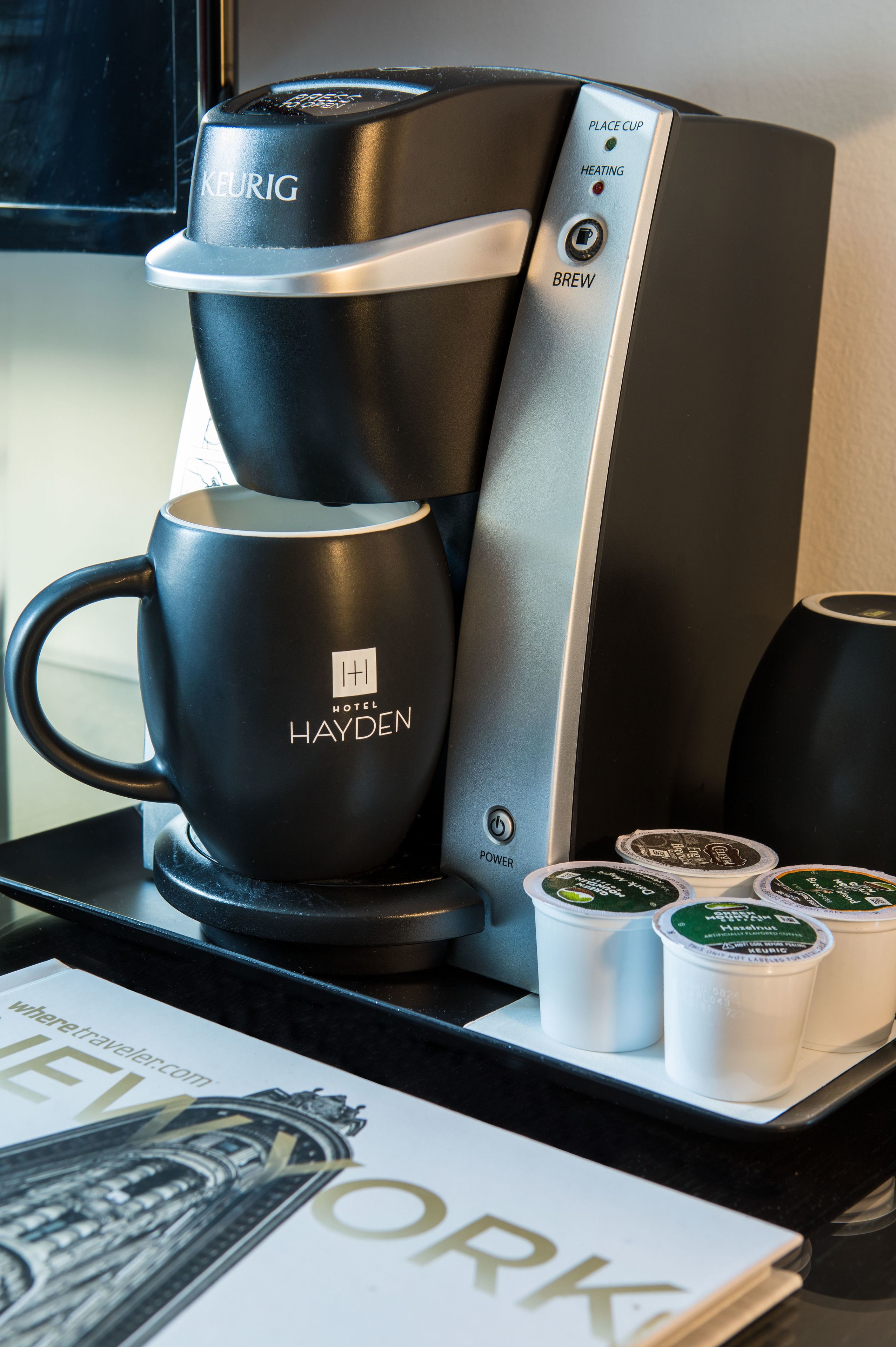 A Keurig coffee machine with a black and silver design, a mug labeled "Hotel Hayden," and three reusable coffee pods on a counter.