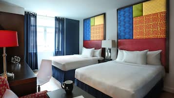 Quarto de hotel com duas camas, uma escrivaninha com luminária, uma cadeira e uma mesa de centro.