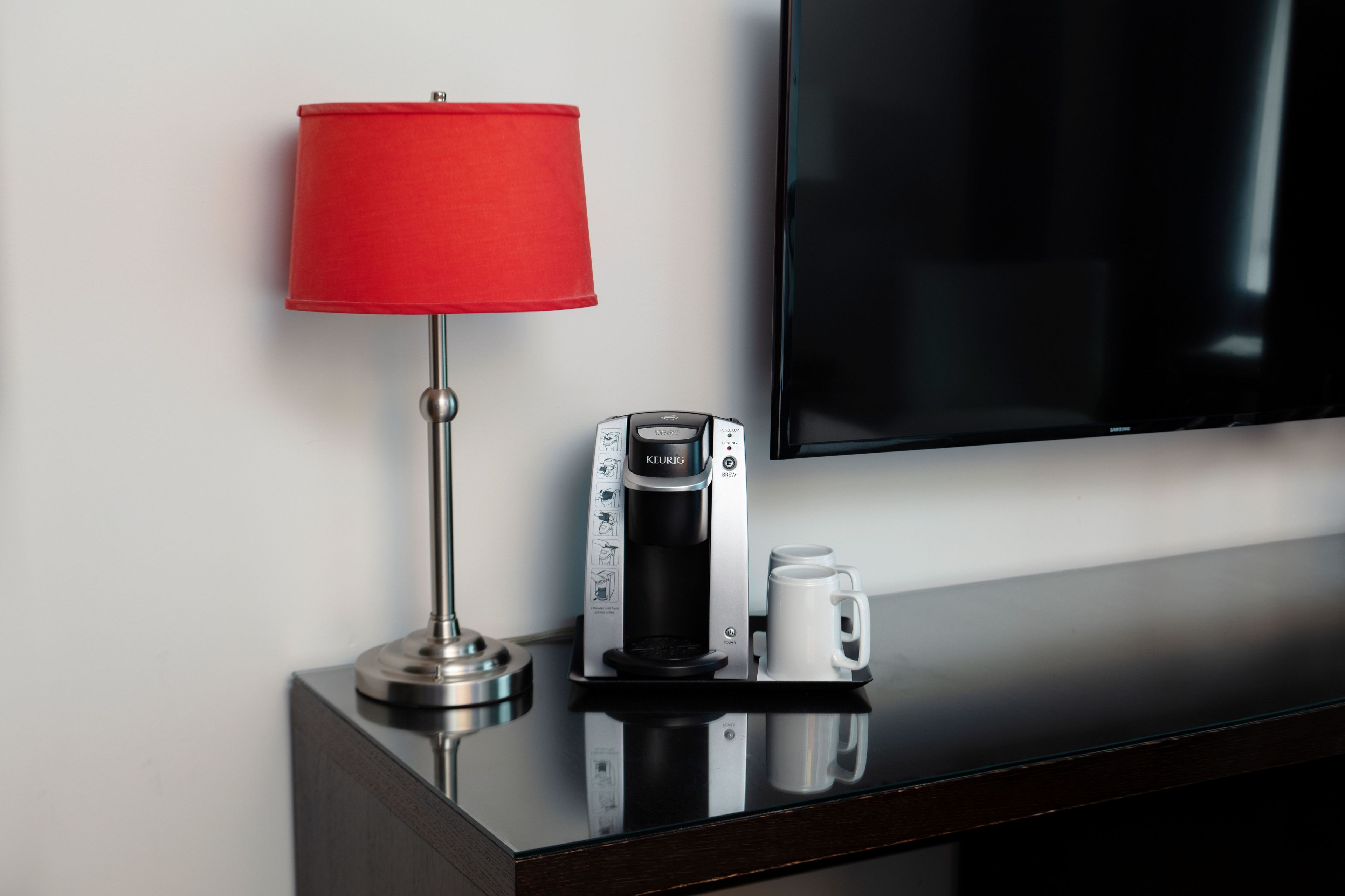 Une chambre d’hôtel moderne, dotée d’un téléviseur noir au design épuré, d’une lampe rouge et d’une machine à café posée sur une surface réfléchissante.