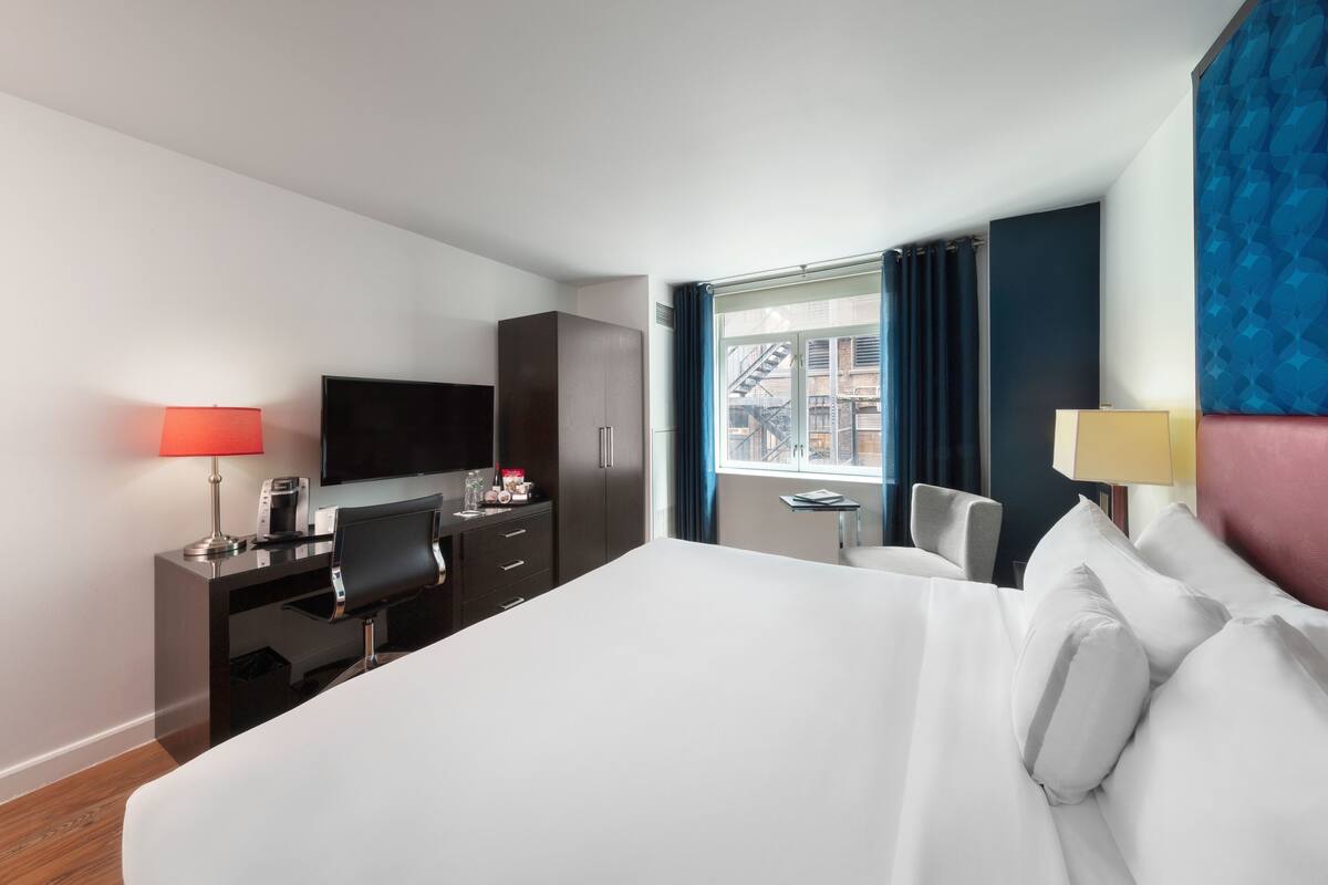A hotel room with a bed, desk, chair, TV, and a window with a view of buildings.