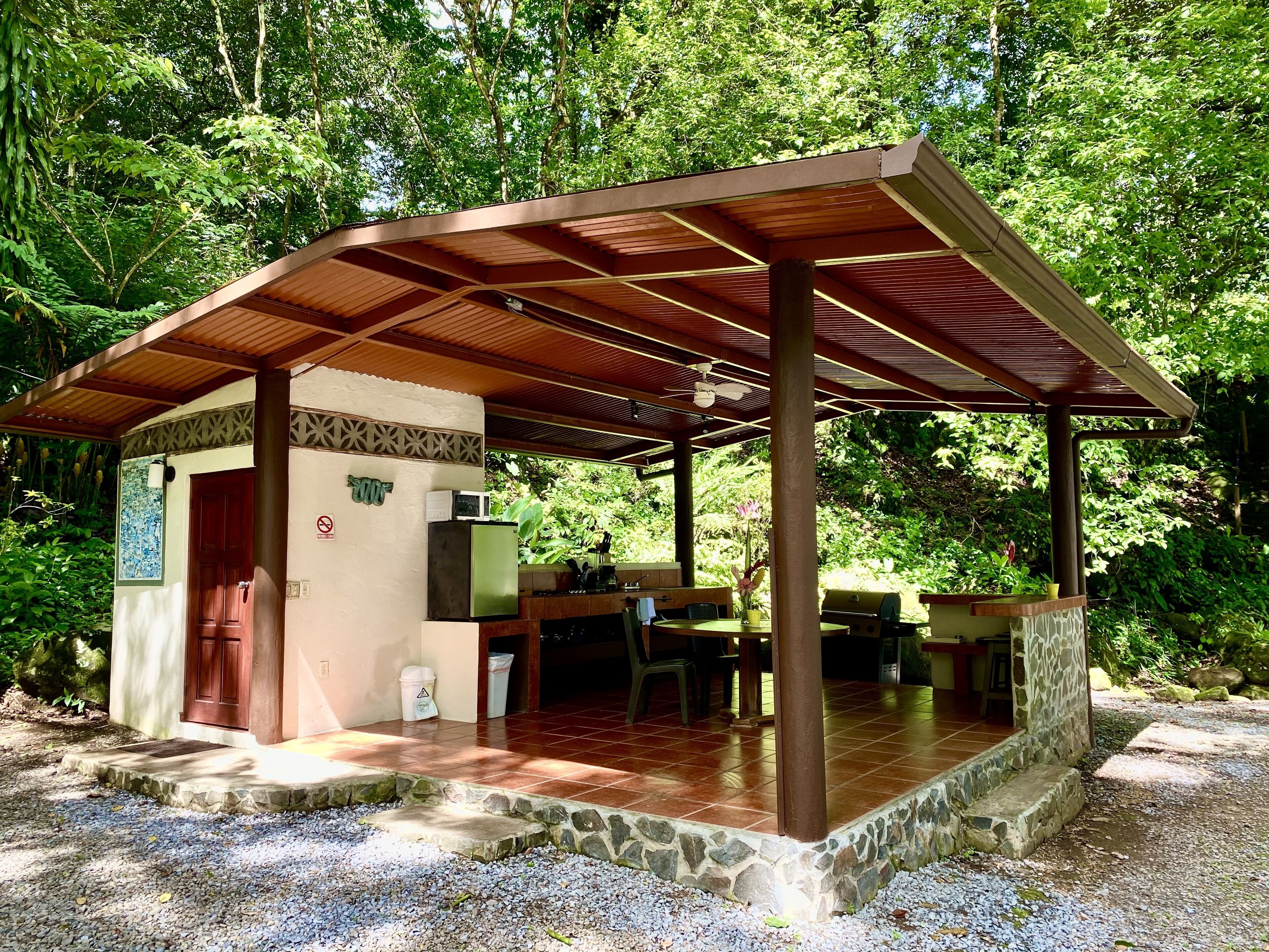 Un espacio exterior techado con una plataforma de piedra, un refrigerador, una parrilla y una mesa de comedor.