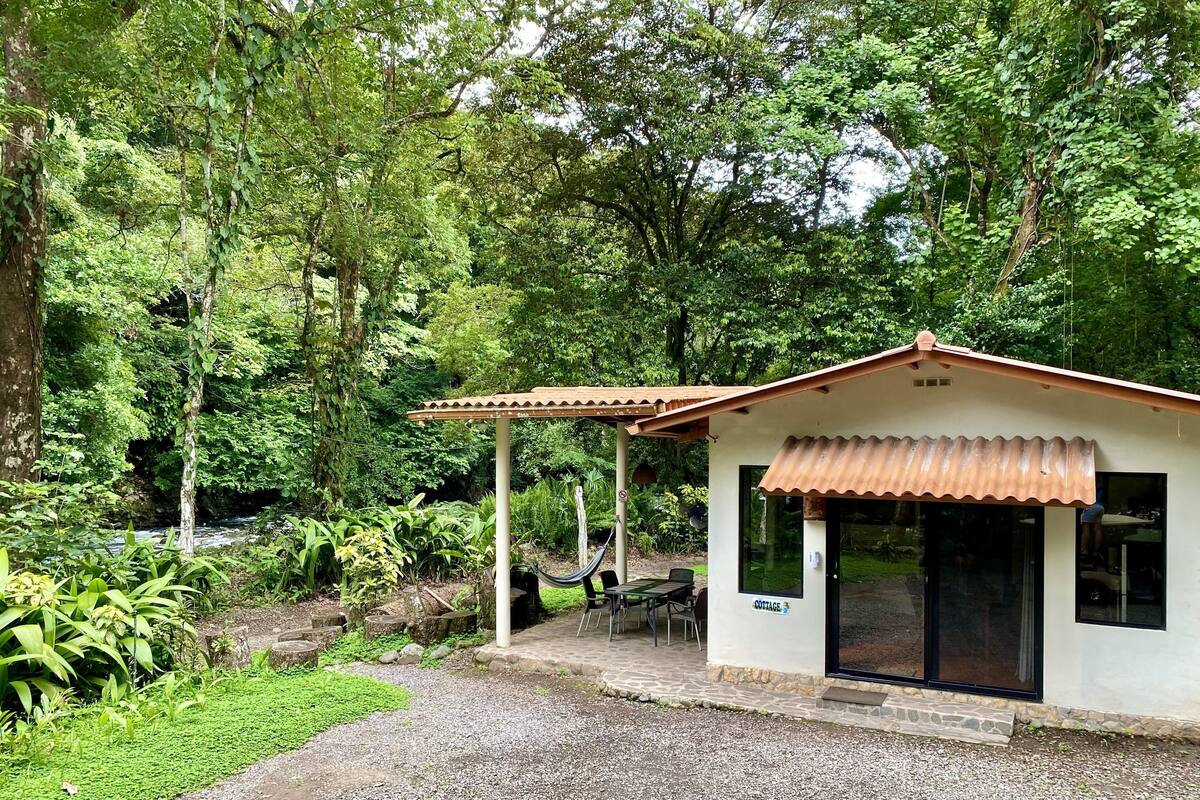 A small building with a tiled roof and a 'COTTAGE' sign, surrounded by lush greenery and a gravel path.